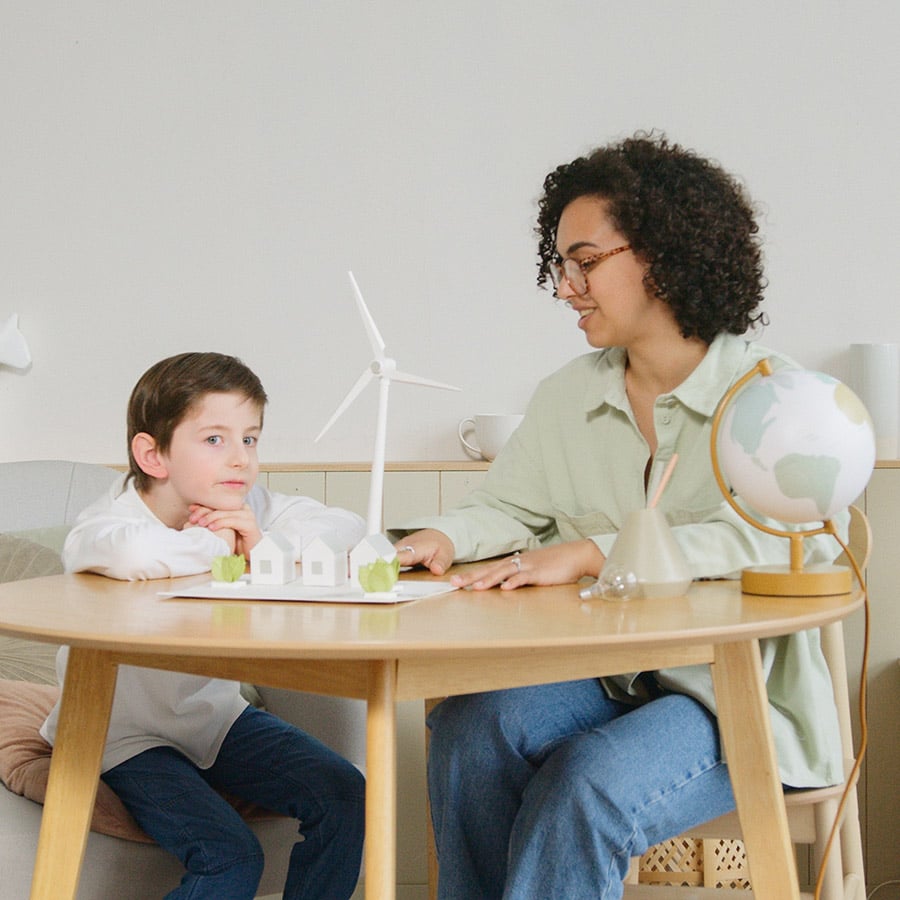 Eine Erzieherin sitzt mit einem Jungen an einem Tisch. Sie schauen sich ein Modell mit Häusern und einem Windrad an. 