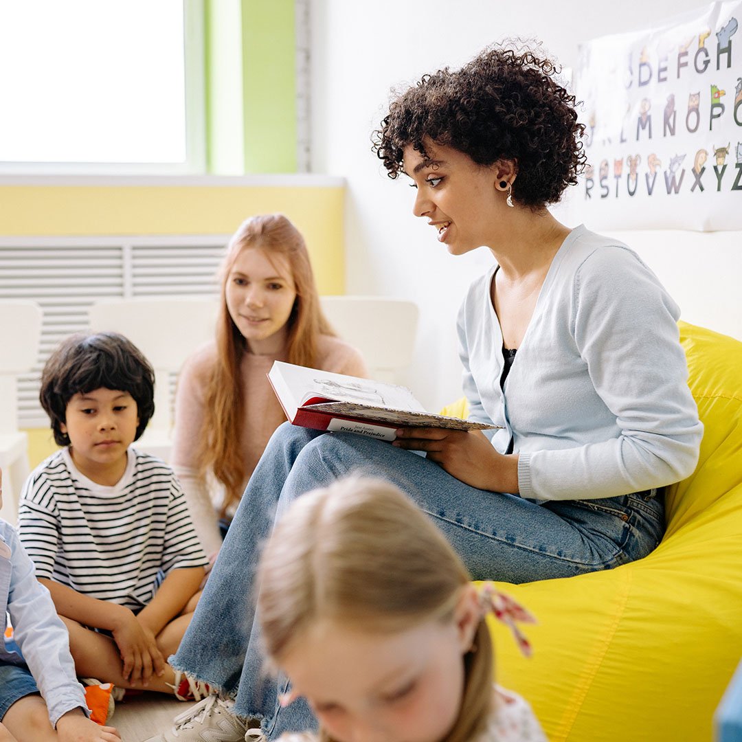 Erzieherin sitzt auf einem gelben Sitzsack und liest drei Kindern aus einem Buch vor. Eine zweite Erzieherin sitzt neben ihr.